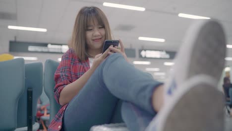 Happy-Asian-woman-using-and-checking-her-smartphone-while-sitting-on-chair-in-terminal-hall-while-waiting-her-flight-at-the-departure-gate-in-international-airport.-Women-happy-in-airport-concept.