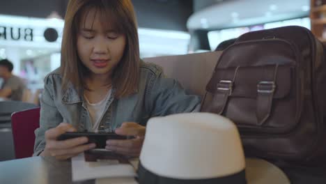 Happy-Asian-woman-using-and-checking-her-smartphone-while-sitting-on-chair-in-terminal-hall-while-waiting-her-flight-at-the-departure-gate-in-international-airport.-Women-happy-in-airport-concept.