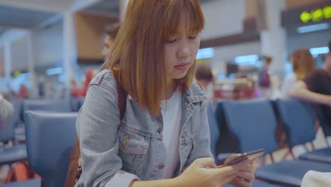 Happy-Asian-woman-using-and-checking-her-smartphone-while-sitting-on-chair-in-terminal-hall-while-waiting-her-flight-at-the-departure-gate-in-international-airport.-Women-happy-in-airport-concept.