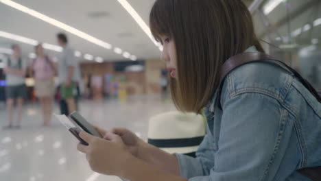 Happy-Asian-woman-using-and-checking-her-smartphone-while-sitting-on-chair-in-terminal-hall-while-waiting-her-flight-at-the-departure-gate-in-international-airport.-Women-happy-in-airport-concept.