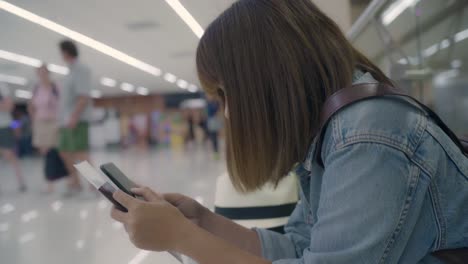 Happy-Asian-woman-using-and-checking-her-smartphone-while-sitting-on-chair-in-terminal-hall-while-waiting-her-flight-at-the-departure-gate-in-international-airport.-Women-happy-in-airport-concept.