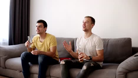 Front-view-of-two-roommates,-friends-watching-football-on-tv-sitting-in-a-comfortable-sofa-in-the-living-room-at-home,-eating-chips-from-the-red-bowl-and-drinking-beer,-celebrating-the-win