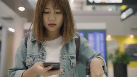 Happy-Asian-woman-using-and-checking-her-smartphone-in-terminal-hall-while-waiting-her-flight-at-the-departure-gate-in-international-airport.-Women-happy-in-airport-concept.