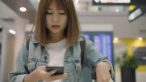 Feliz-mujer-asiática-utilizando-y-controlando-su-smartphone-en-el-hall-de-la-terminal-mientras-espera-su-vuelo-en-la-puerta-de-salida-en-el-aeropuerto-internacional.-Mujer-feliz-en-concepto-de-aeropuerto.