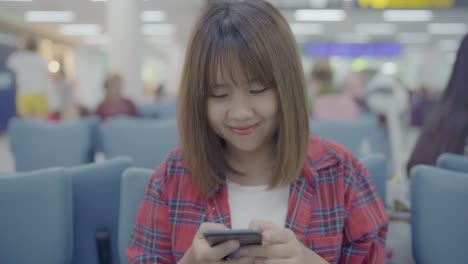 Happy-Asian-woman-using-and-checking-her-smartphone-while-sitting-on-chair-in-terminal-hall-while-waiting-her-flight-at-the-departure-gate-in-international-airport.-Women-happy-in-airport-concept.