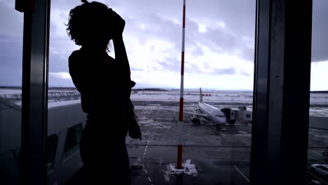 Niña-de-pie-en-el-aeropuerto-cerca-de-una-ventana-grande-mirando-los-planos-en-la-pista.-Silueta-de-una-joven-contra-el-cielo-gris-y-el-pie-en-el-avión-de-territorio