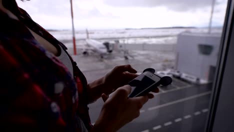 Muchacha-en-camisa-a-cuadros-ve-mensajes-de-amigos,-a-continuación,-comprueba-el-correo-electrónico-en-el-aeropuerto-contra-la-ventana-en-la-que-el-plano-visible-y-un-campo-cubierto-de-nieve