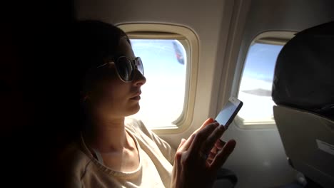 Tourist-woman-sitting-near-airplane-window-at-sunset-and-using-mobile-phone-during-flight