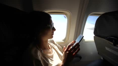 Mujer-turista-sentada-junto-a-la-ventana-del-avión-al-atardecer-y-usar-teléfono-celular-durante-el-vuelo