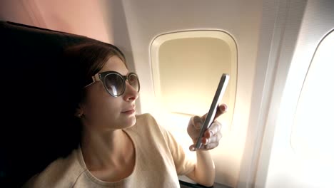 Tourist-woman-sitting-near-airplane-window-at-sunset-and-using-mobile-phone-during-flight