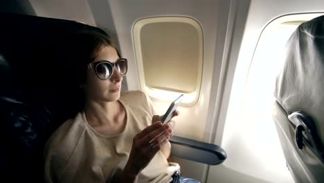 Tourist-woman-sitting-near-airplane-window-at-sunset-and-using-mobile-phone-during-flight