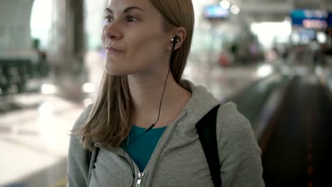 Happy-smiling-woman-using-travolator-in-airport-terminal.-Using-her-smartphone,-listening-to-music