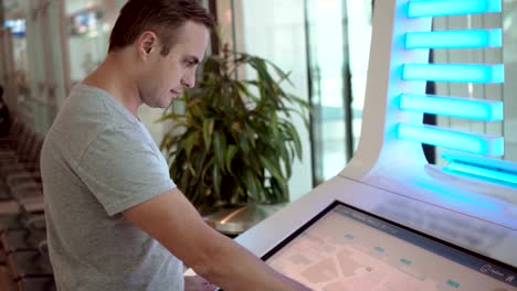 Handsome-guy-in-grey-t-shirt-in-airport-terminal-using-touchscreen-searching-for-his-flight-information