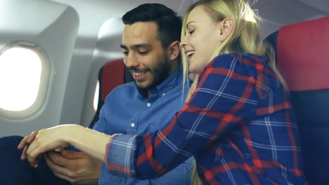 Bei-einem-Board-von-kommerziellen-Flugzeug-schöne-junge-Blondine-mit-schönen-hispanische-männlich-spielen-auf-Smartphone-und-Lächeln.-Durch-Flugzeug-Fenster-scheint-die-Sonne.