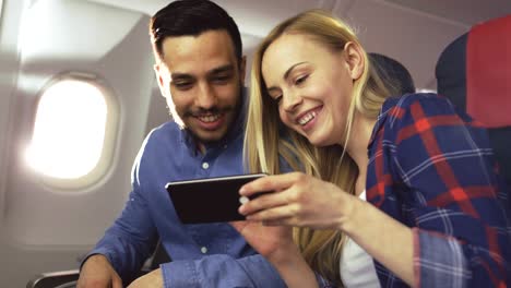 Bei-einem-Board-von-kommerziellen-Flugzeug-schöne-junge-Blondine-mit-schönen-hispanische-männlich-teilen-Sie-Videos-auf-Smartphone-und-Lächeln.-Durch-Flugzeug-Fenster-scheint-die-Sonne.