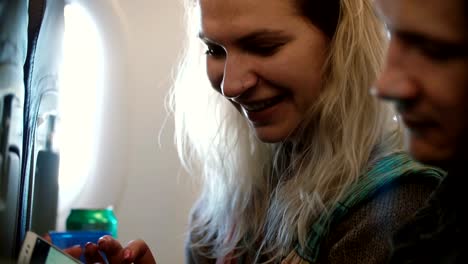 Young-beautiful-couple-traveling-by-plane.-Blonde-woman-and-man-sitting-near-the-window-and-using-smartphone