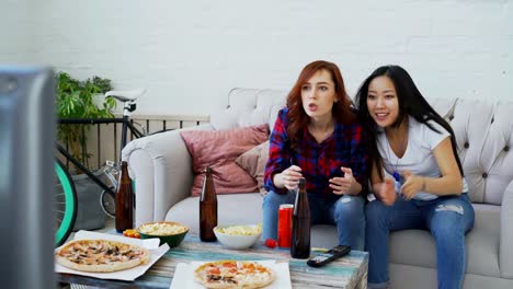 Dos-amigas-multiétnicas-viendo-deportes-partido-en-TV-juntos-en-casa-bebiendo-cerveza-y-comiendo-snacks