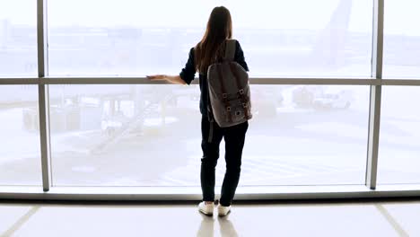 Young-woman-with-backpack-near-terminal-window.-Caucasian-female-tourist-using-smartphone-in-airport-lounge.-Travel.-4K