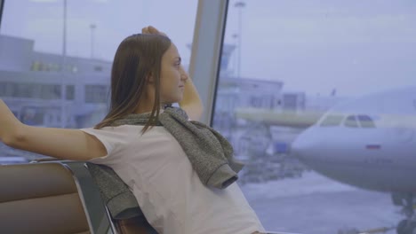 Young-woman-sits-and-stretches-at-airport-with-airplane-on-the-background