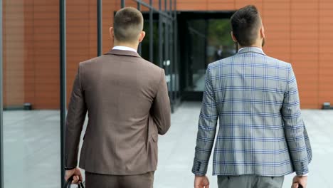Two-businessmen-chat-together-as-they-walk-along-through-a-busy-modern-office-building.-Rear-view
