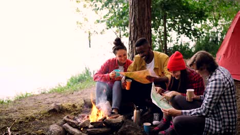 Lazo-del-Cinemagraph---sonreír-a-chicos-y-chicas-mirando-mapas-de-descanso-alrededor-del-fuego-en-bosques-y-tenencia-de-bebidas.-Llama-y-el-papel-se-mueve,-hermosa-naturaleza-es-visible.