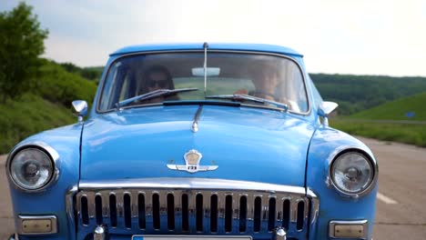 Young-couple-in-hats-riding-in-vintage-car-at-summer-travel.-Man-and-woman-sitting-at-the-front-seat-of-old-retro-car-during-ride.-Concept-of-road-trip-at-holidays.-Slow-motion-Close-up-Front-view