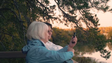 Two-elderly-woman-looking-smartphone-at-autumn-walk-on-tree-and-lake-landscape