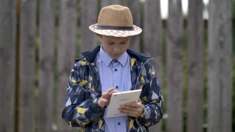 farmer-boy-in-a-hat-uses-a-tablet,-stands-against-the-fence