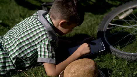 niño-se-encuentra-en-la-hierba-con-una-tableta-después-de-un-paseo-en-bicicleta,-al-aire-libre