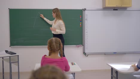 Junior-School,-mujer-del-profesor-dibuja-figuras-geométricas-a-bordo-durante-la-lección-para-los-escolares-sentados-en-pupitres-en-el-aula