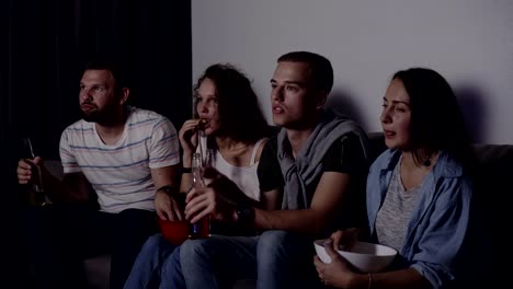 Freunde-von-vier-sitzen-auf-der-Couch,-Film-ansehen-oder-im-Fernsehen-zu-zeigen.-Kaukasische-junge-Gruppe-sitzen-auf-der-Couch,-trinken-Bier-und-Essen-Popcorn-aus-der-roten-Schale.-Film-Nacht