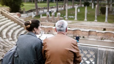 Rückansicht-des-Europäischen-Mädchen-und-ältere-Mann-sitzen-und-reden-an-alten-Ruinen-der-antiken-Ostia-Amphitheater-Italiens.