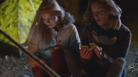 Two-happy-girls-are-sitting-next-to-a-campfire-at-night-and-use-their-smartphones.