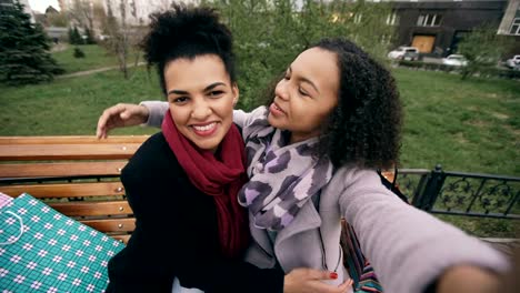 Dos-linda-mujer-afroamericana-tomando-selfie-en-smartphone-con-bolsas-de-compras-y-sonriente.-Sentado-en-el-Banco-de-la-calle-novias-divertirse-después-de-visitar-el-centro-comercial-ventas