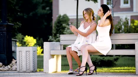 Cute-and-adult-girls-who-are-dressed-in-summer-clothes-do-selfie-in-the-park,-the-ladies-sit-on-a-bench-on-a-warm-day