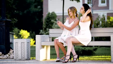 Lindas-y-adultos-chicas-que-se-visten-con-ropa-de-verano-hacen-selfie-en-el-parque,-el-señoras-se-sientan-en-un-banco-en-un-día-caluroso