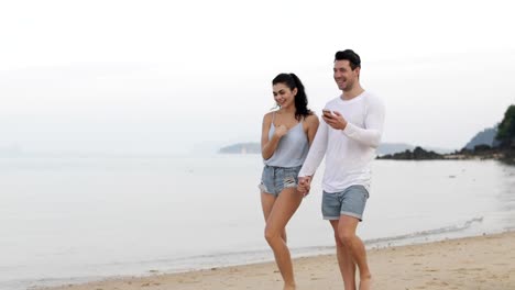 Pareja-caminando-en-la-playa-con-teléfonos-inteligentes-hablar,-hombre-joven-y-los-turistas-mujer-cogidos-de-la-mano