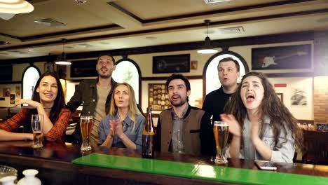 Students-sports-fans-are-watching-championship-in-bar-while-sitting-at-counter-and-drinking-beer.-They-are-cheering-anxiously,-then-celebrating-success.