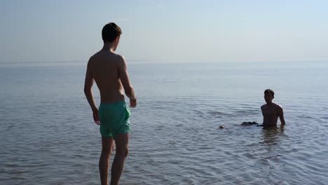 Two-young-men-in-shallow-water,-one-walks-out-of-the-water