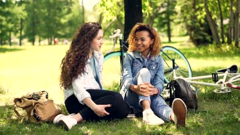 Alegres-estudiantes-afroamericanos-y-caucásicos-están-hablando-y-riendo-sentado-en-el-parque-en-el-césped-después-de-montar-en-bici.-Concepto-naturaleza,-conversación-y-amistad.