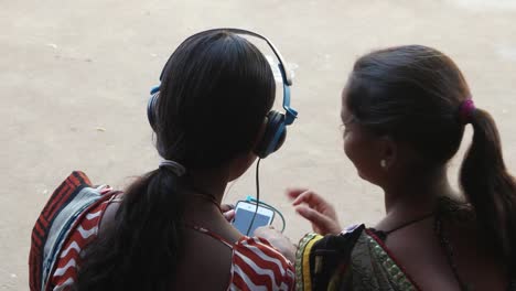 back-of-two-females-sharing-wireless-music-on-phone-mobile-smile-laugh-enjoying-sharing-listening-headphones-together-bonding-love-care-home-interior-close-up-shaking-head-tapping-feet-taking-giving