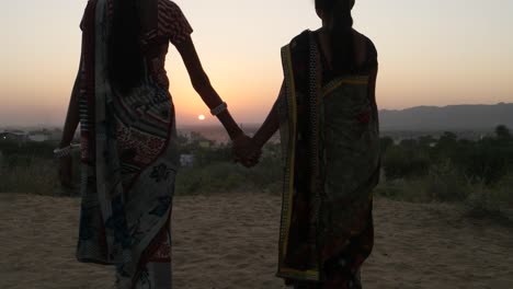 Two-women-stand-hold-hands-at-sunset-beautiful-vantage-point-romantic-setting-dawn-high-wide-panoramic-surreal-lovers-in-traditional-dress-silhouette-in-Rajasthan-India-handheld-from-behind-medium-two