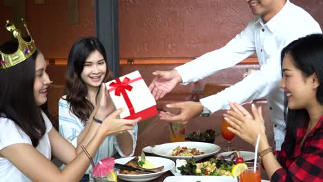Joven-pareja-asiática-dando-una-presente-caja-de-regalo-y-amigos-aplaudiendo-las-manos-cerca-de-restaurante