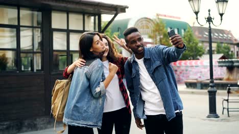 Happy-African-American-guy-is-taking-selfie-with-friends-beautiful-Caucasian-girls-standing-in-street-posing-and-holding-smartphone-during-journey-in-foreign-country.