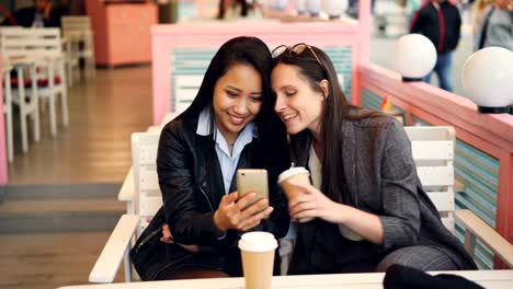 Fröhliche-Junge-Frauen-sind-im-Café-entspannen-und-mit-Smartphone-Bildschirm-schauen-und-Lächeln-auf-den-Lippen-halten-Kaffee-zum-Mitnehmen.-Jugend,-Technik-und-Getränke-Konzept.