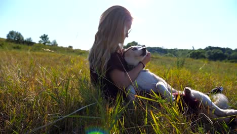 Dolly-shot-of-young-girl-with-blonde-hair-sitting-on-grass-at-field-and-hugging-her-husky-dog.-Beautiful-woman-in-sunglasses-caress-and-kissing-her-pet-at-meadow.-Friendship-with-domestic-animal