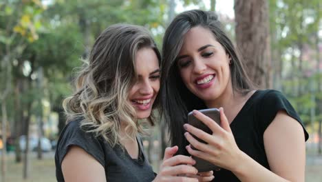 Two-girlfriends-holding-cellphone-laughing-and-smiling-together.-Latin-women-holding-smartphone-laugh-at-content-they-are-watching