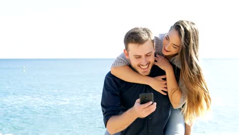 Glückliches-Paar-scherzen-und-mit-Handy-am-Strand