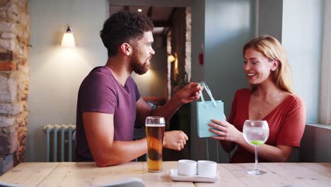 Young-adult-mixed-race-man-giving-a-present-to-his-girlfriend,-sitting-with-drinks-at-a-table-in-a-pub,-close-up,-side-view