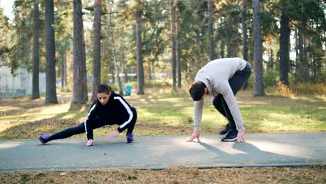 Hombre-guapo-joven-y-amiga-están-entrenando-en-el-Parque-juntos,-estirar-las-piernas-en-cálido-día-de-otoño-con-prendas-de-moda.-Concepto-personas,-deporte-y-ocio.